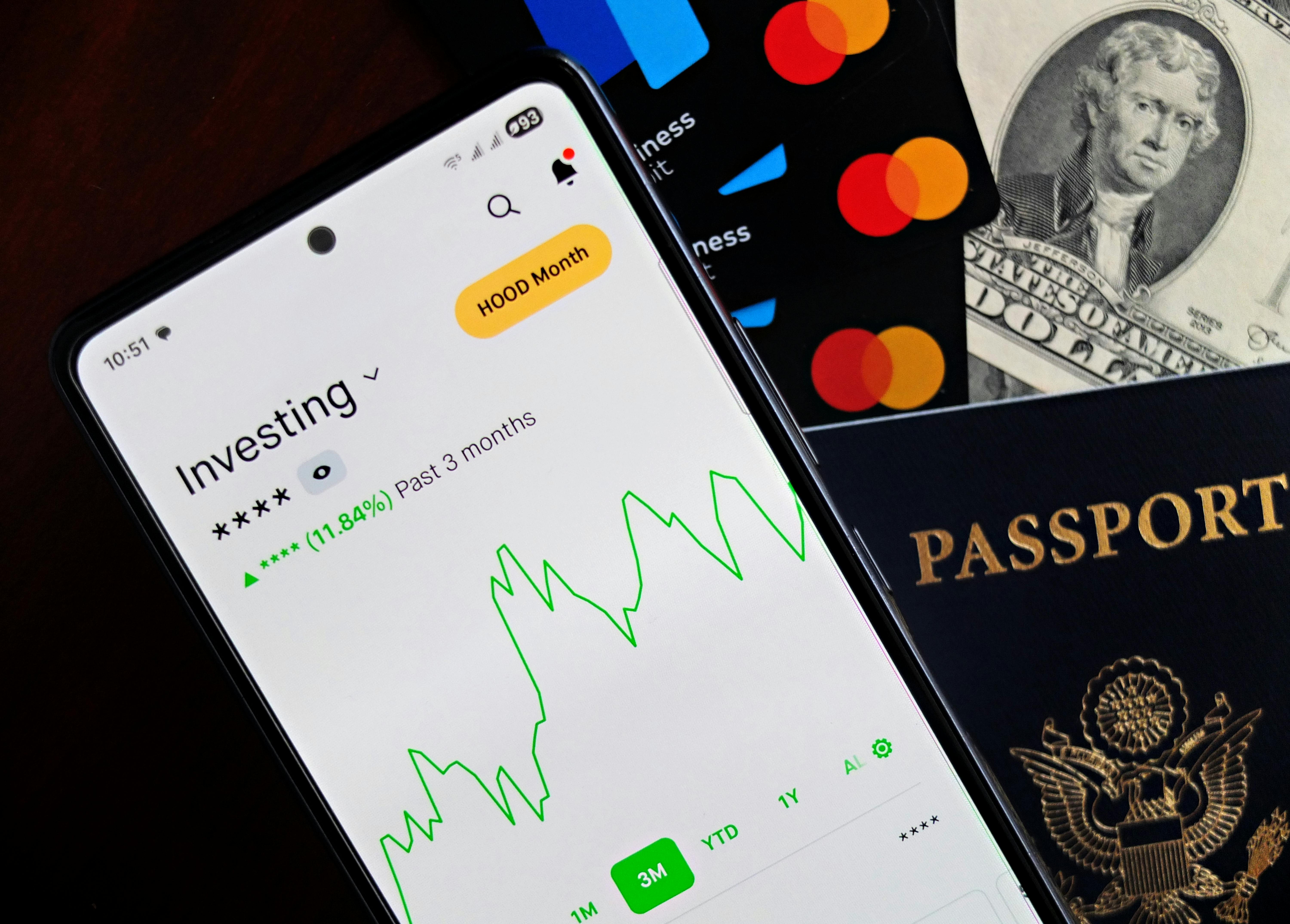 Smartphone displaying investing app with credit cards and cash on a desk, personal finance concept