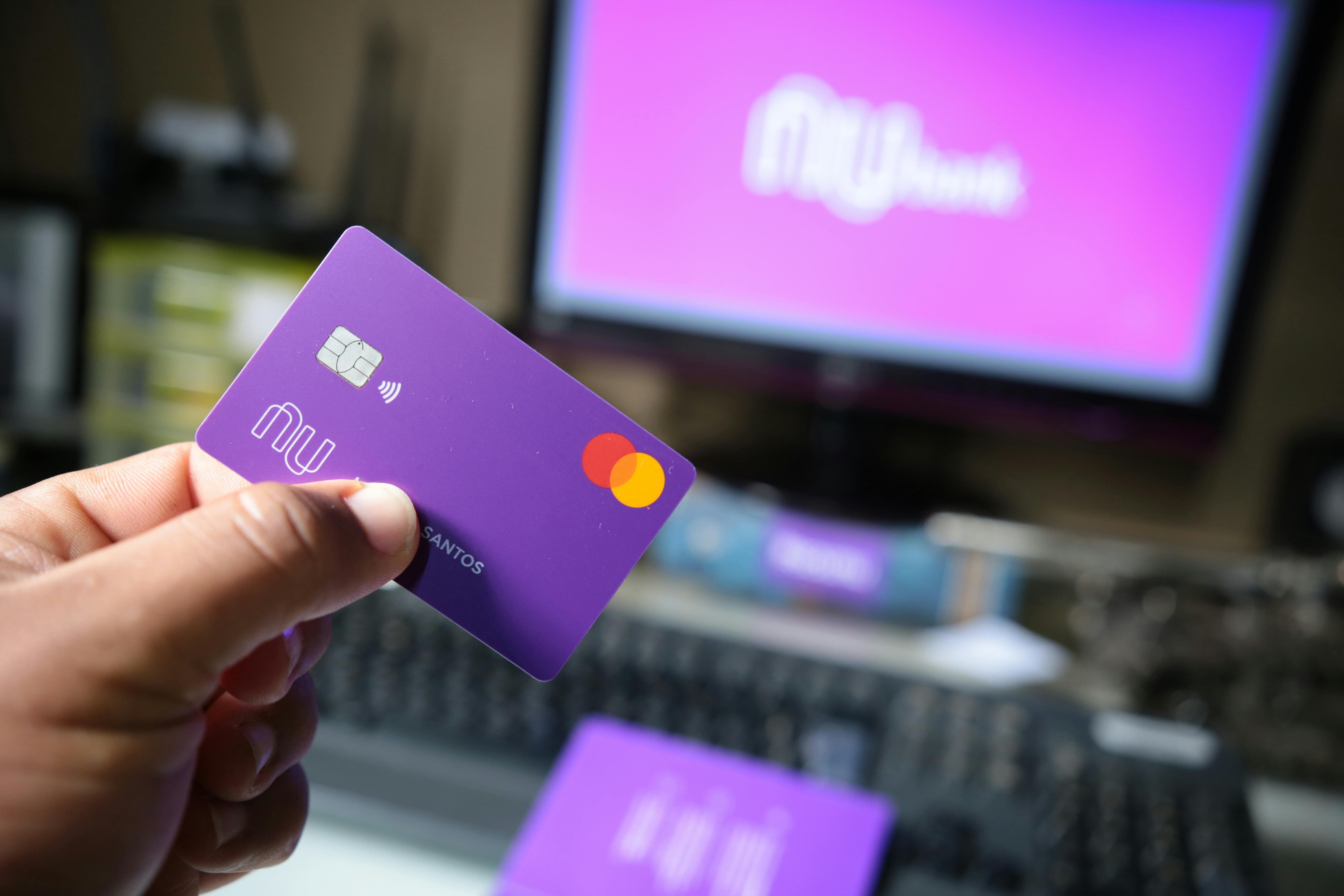Hand holding a purple bank card in front of a computer screen, representing Nubank digital banking