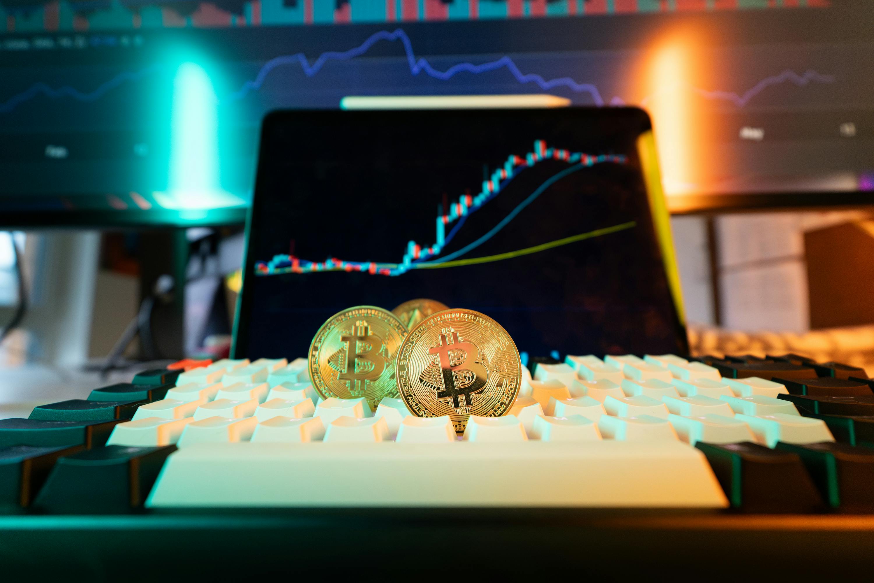 Bitcoin coins on a keyboard with trading charts displayed on monitors in the background