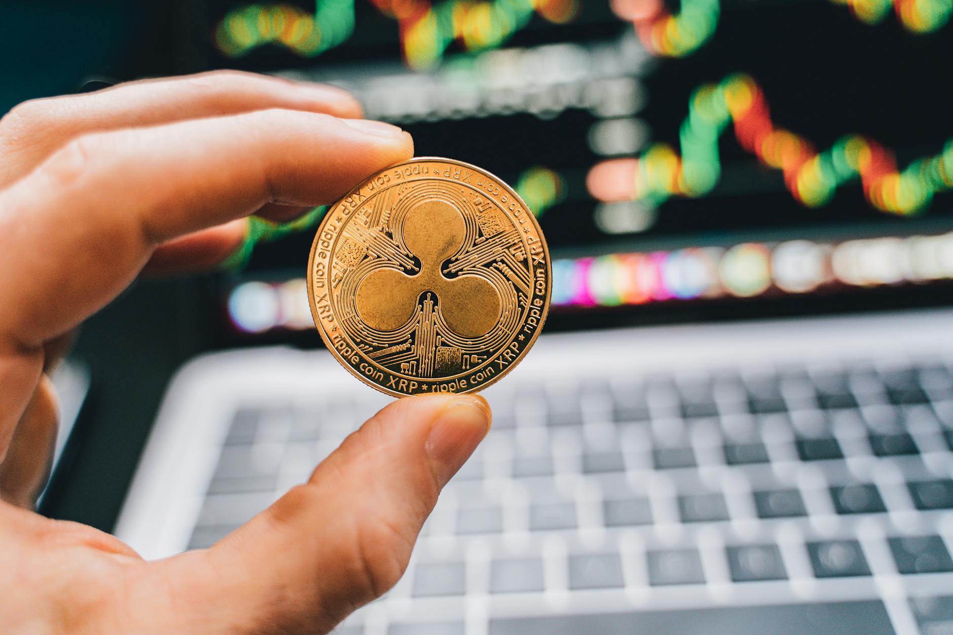 Hand holding a Ripple XRP coin with a computer screen blurred in the background