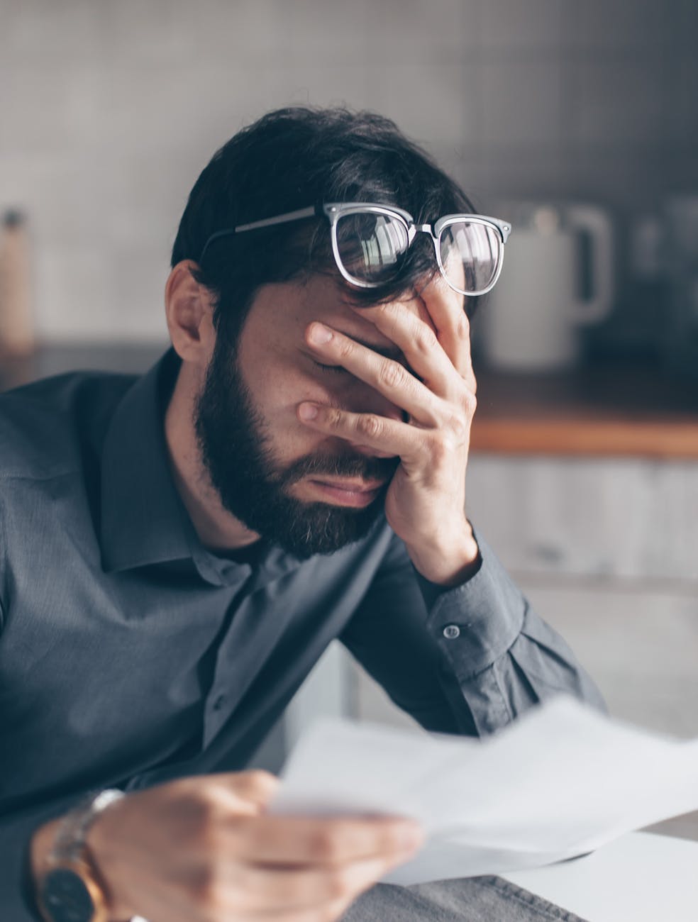 Man with glasses holding head in frustration while reading financial documents, indicating financial stress