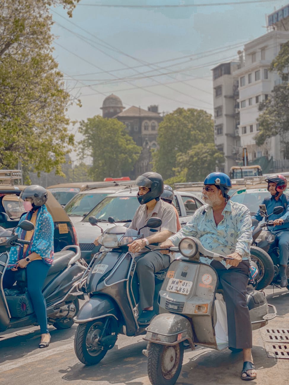 Crowded street in Mumbai with adults riding scooters amidst traffic and buildings.