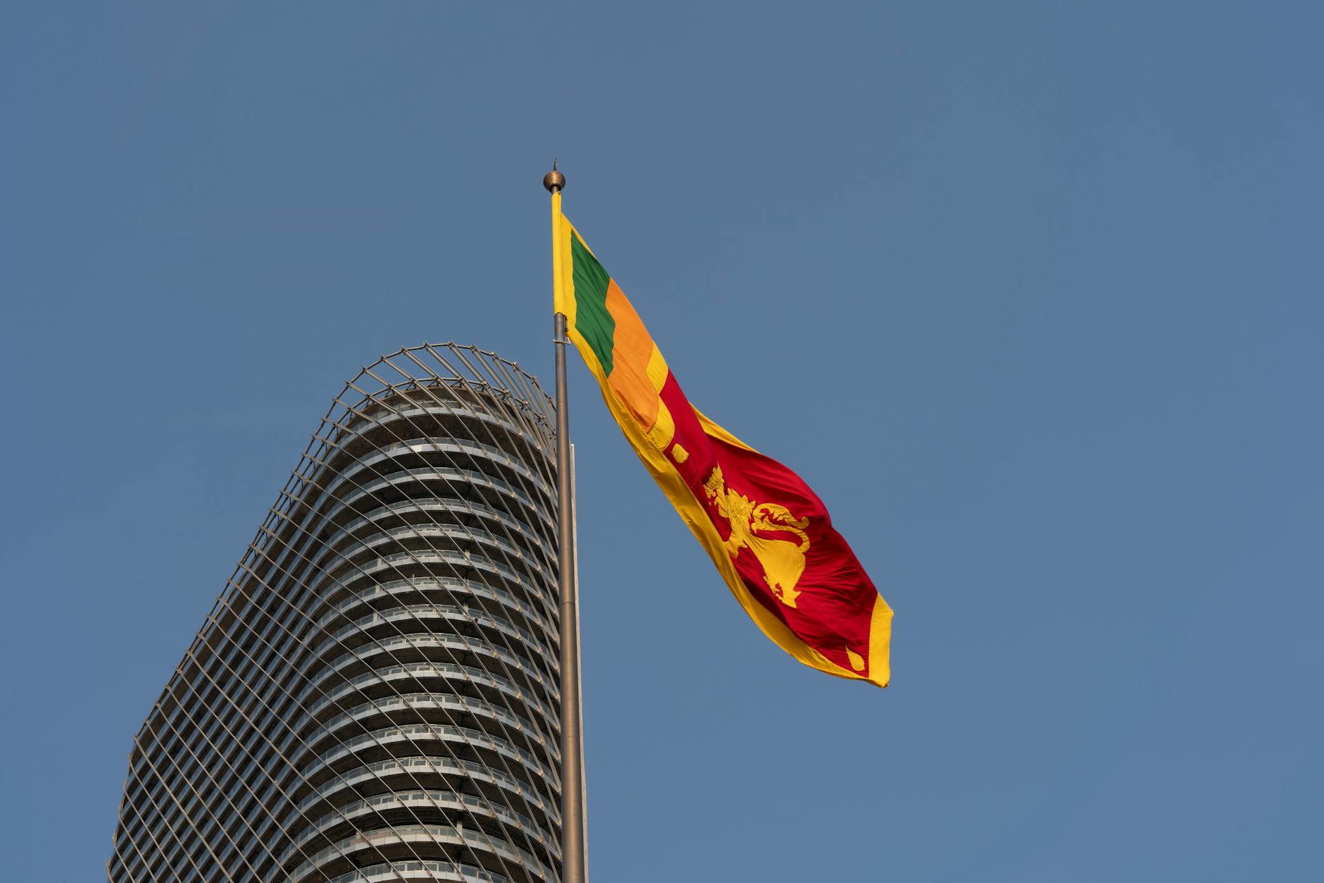 The Sri Lankan flag waving beside a modern Colombo skyscraper against a clear blue sky.